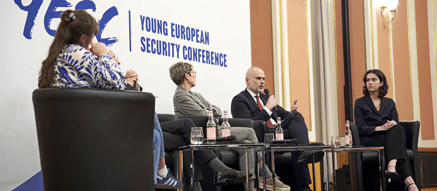 Vier Personen sitzen auf Sesseln vor einer Wand mit der Aufschrift 'YOUNG EUROPEAN SECURITY CONFERENCE'