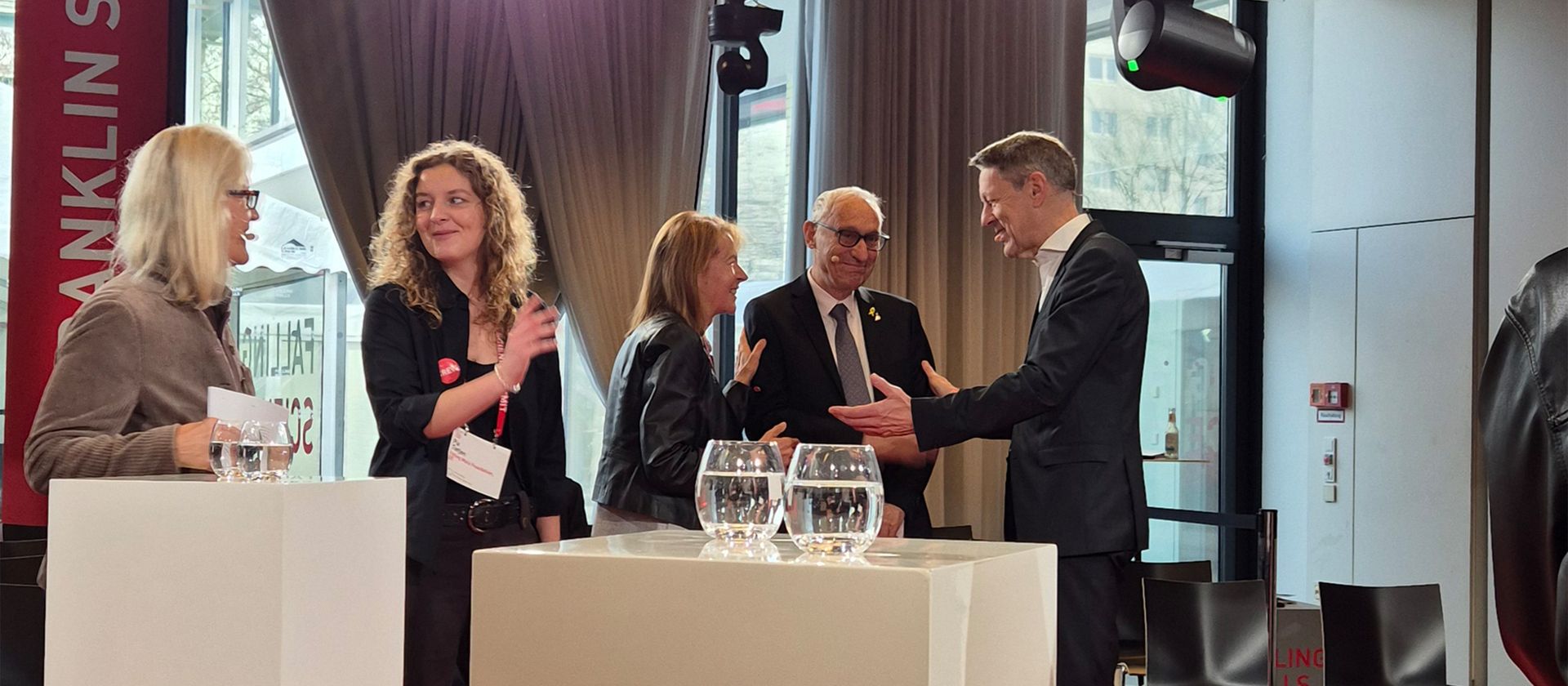 Fünf Personen in formeller Kleidung stehen in einem Raum mit großen Fenstern und Vorhängen und unterhalten sich um einen weißen Stehtisch mit zwei Wasserkaraffen