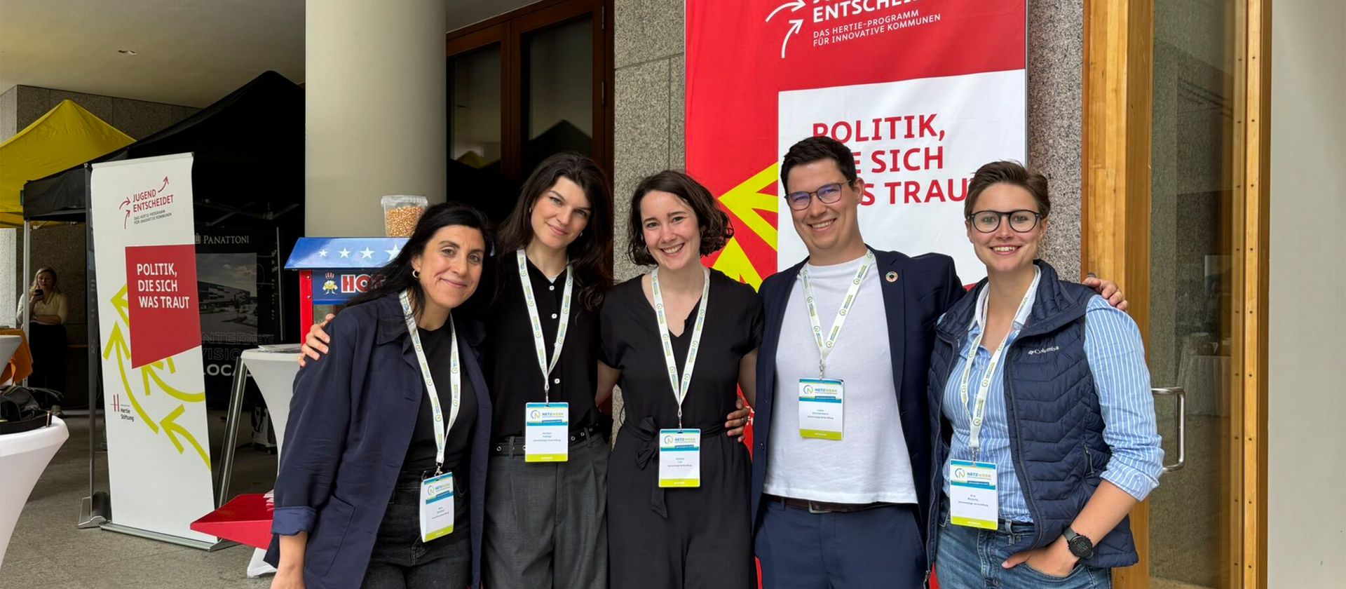 Fünf Personen mit Namensschildern stehen vor einem roten Banner mit dem Text 'POLITIK, DIE SICH WAS TRAUT' und posieren für ein Gruppenfoto