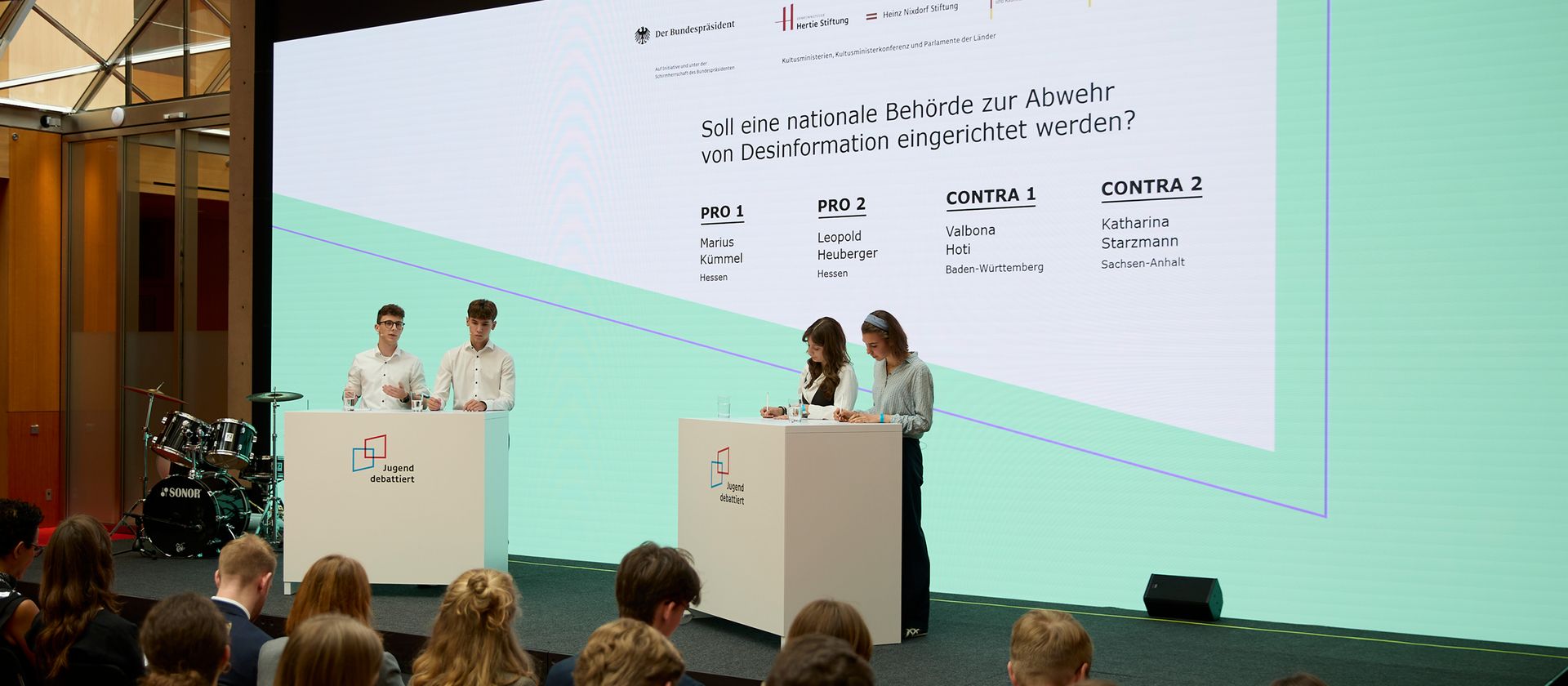 Zwei Gruppen Jugendlicher debattieren an zwei Pulten vor Publikum und großer Projektionswand mit Text zu nationaler Behörde gegen Desinformation