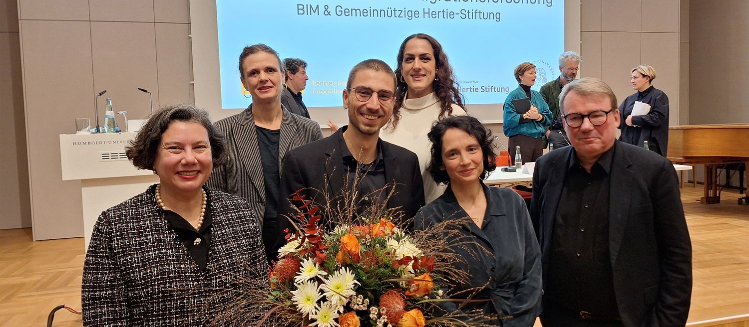 Gruppe von sechs Personen steht vor einer Wand mit Text, eine Person hält einen großen Blumenstrauß