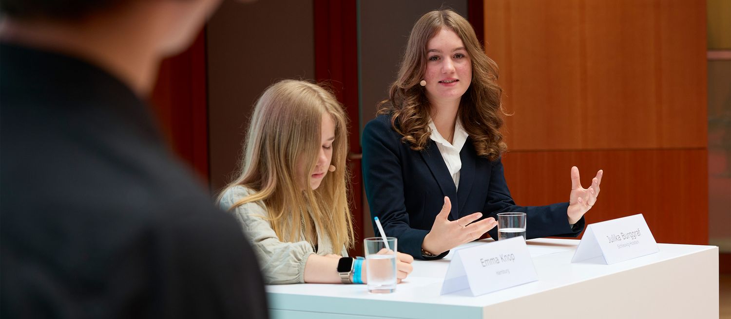 Zwei junge Frauen sitzen an einem Tisch mit Namensschildern und sprechen, eine gestikuliert mit beiden Händen