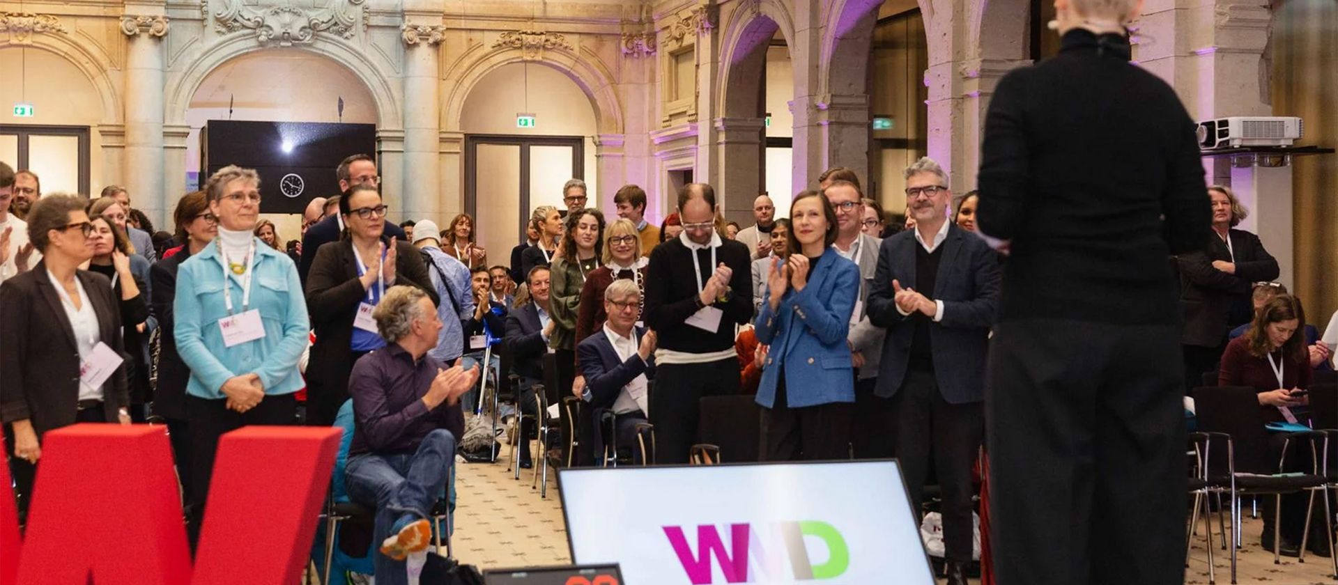 Menschen in einem großen Saal applaudieren einer Person im Vordergrund, die mit dem Rücken zur Kamera steht