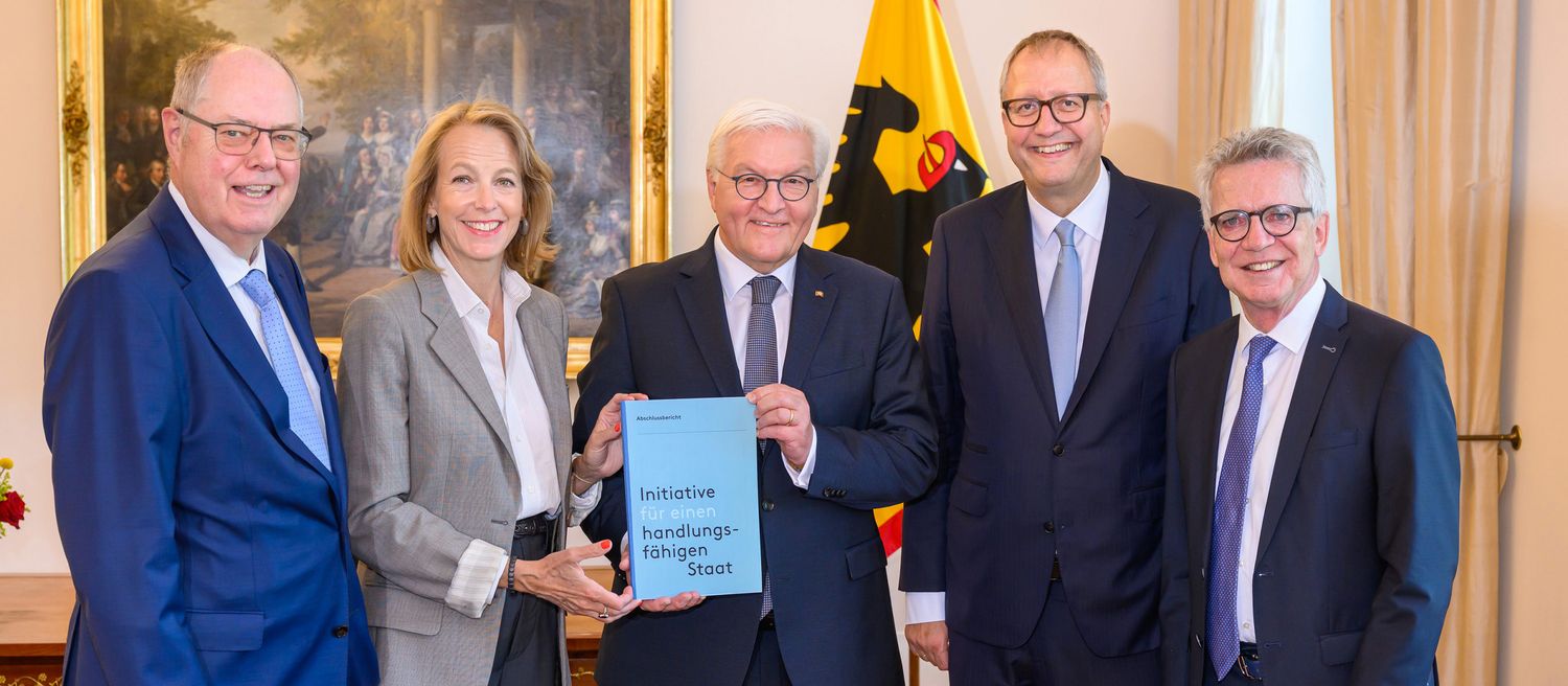 Fünf Personen in Anzügen stehen in einem Raum, eine Person hält ein hellblaues Dokument mit der Aufschrift 'Initiative handlungsfähiger Staat'