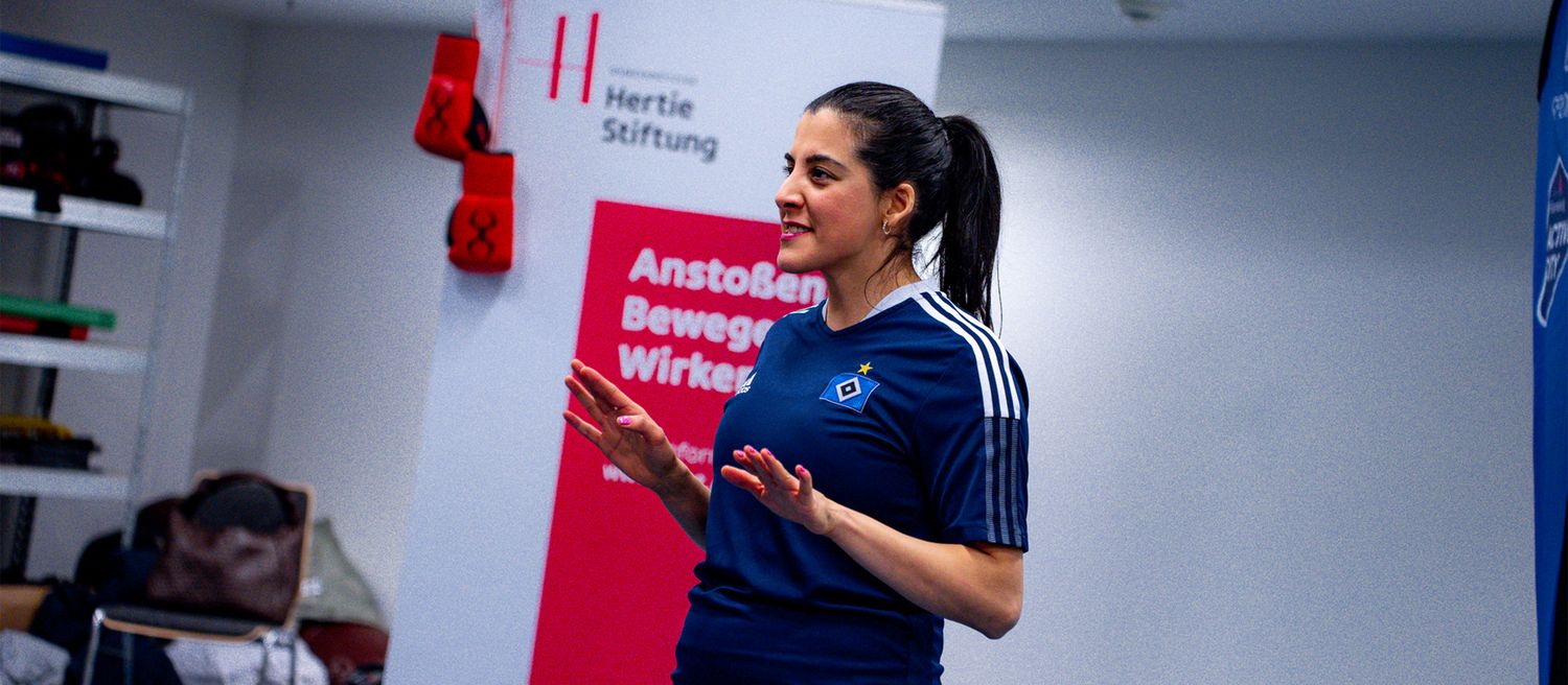 Frau mit Pferdeschwanz in blauem Sportshirt mit weißem Logo und drei weißen Streifen an den Ärmeln gestikuliert vor einem Banner mit rotem und weißem Design