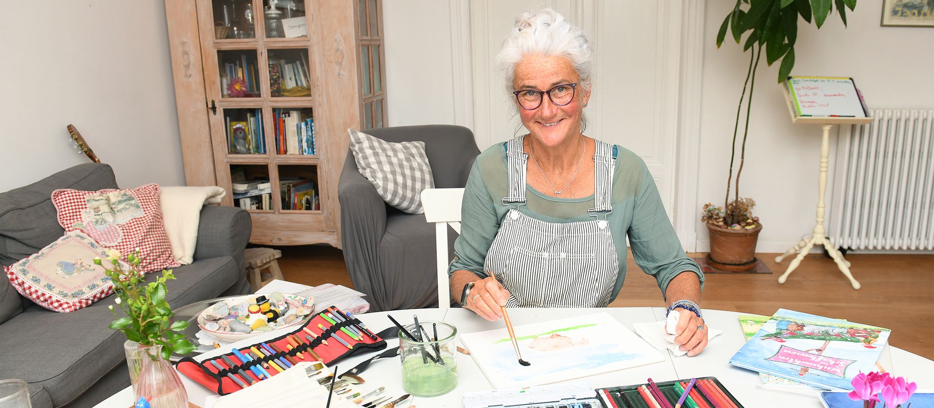 Ältere Frau mit grauem Haar und Schürze sitzt an einem Tisch und malt mit Pinsel auf Papier in einem Wohnzimmer mit Sofa, Bücherregal und Pflanzen