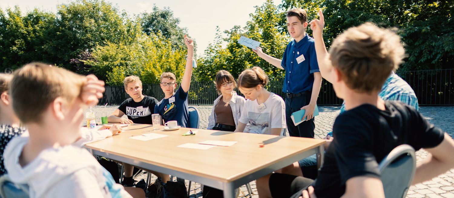 Gruppe von Jugendlichen sitzt an einem Tisch im Freien, zwei Jugendliche heben die Hand, eine Person steht mit Notizblock