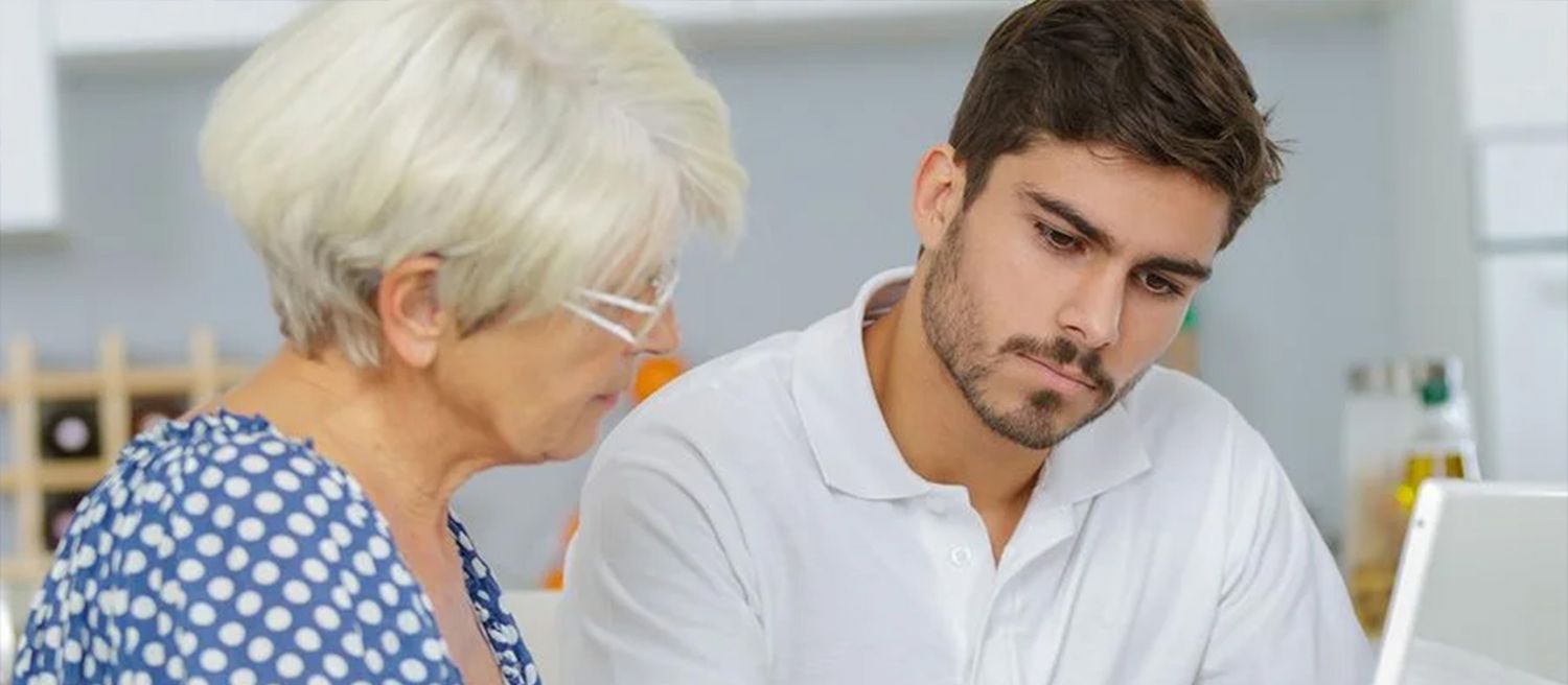 Ältere Frau mit weißem kurzen Haar und blau-weiß gepunkteter Bluse im Gespräch mit jüngeren Mann in weißem Poloshirt in Innenraum